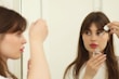A woman with brown hair is applying skincare serum to her face using a dropper. She is holding the dropper above her cheek near a mirror, and her reflection is visible. She appears to be in a well-lit room, wearing a white top.