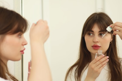 A woman with brown hair is applying skincare serum to her face using a dropper. She is holding the dropper above her cheek near a mirror, and her reflection is visible. She appears to be in a well-lit room, wearing a white top.