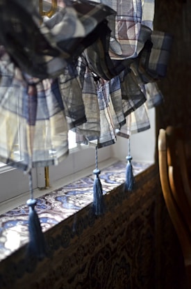 A close-up of a windowsill adorned with intricately patterned tiles and a sheer, plaid valance with blue tassels hanging down. The windowsill and valance create a cozy and somewhat vintage atmosphere. The light filters softly through the sheer fabric of the valance, adding a gentle and warm ambiance to the scene.