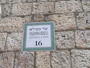 A street sign mounted on a textured stone wall displays text in three languages: Hebrew, Arabic, and English. It indicates the street name as Mamilla Avenue and the number 16.