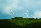 Two wind turbines stand prominently against a backdrop of lush green hills and a partly cloudy sky. The turbines, with their tall white towers and red-tipped blades, are positioned on the crest of the hill, capturing the wind with their large rotors.