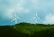 Two wind turbines stand prominently against a backdrop of lush green hills and a partly cloudy sky. The turbines, with their tall white towers and red-tipped blades, are positioned on the crest of the hill, capturing the wind with their large rotors.