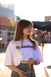 A young woman stands outdoors, wearing a white t-shirt with 'JIL SANDER' printed on it and a floral-patterned skirt. She holds sunglasses in one hand and has long brown hair. The background is bright and sunlit, with blurred buildings and signs.