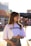 A young woman stands outdoors, wearing a white t-shirt with 'JIL SANDER' printed on it and a floral-patterned skirt. She holds sunglasses in one hand and has long brown hair. The background is bright and sunlit, with blurred buildings and signs.