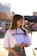 A young woman stands outdoors, wearing a white t-shirt with 'JIL SANDER' printed on it and a floral-patterned skirt. She holds sunglasses in one hand and has long brown hair. The background is bright and sunlit, with blurred buildings and signs.