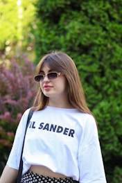 A young woman with long brown hair is wearing sunglasses and a white T-shirt with text on it. She is standing outdoors in front of a lush green background, with some purple foliage visible on the left side.