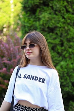 A young woman with long brown hair is wearing sunglasses and a white T-shirt with text on it. She is standing outdoors in front of a lush green background, with some purple foliage visible on the left side.