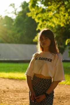 A young woman stands outdoors during a sunny day. She is wearing a white crop top with the text 'JL SANDER' and a black skirt with small floral patterns. Her long hair falls down naturally as she gazes towards the camera. The sunlight creates a warm halo around her, with lush green trees and foliage in the background.