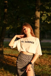 A woman stands in a sunlit forest area, wearing a white shirt with 'JIL SANDER' printed on it and a floral skirt. She is gently touching her neck with one hand and holds sunglasses in the other. An electronic watch is on her wrist. The sunlight casts a warm glow on her face and attire.