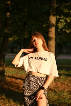A woman stands in a sunlit forest area, wearing a white shirt with 'JIL SANDER' printed on it and a floral skirt. She is gently touching her neck with one hand and holds sunglasses in the other. An electronic watch is on her wrist. The sunlight casts a warm glow on her face and attire.