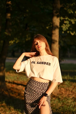 A woman stands in a sunlit forest area, wearing a white shirt with 'JIL SANDER' printed on it and a floral skirt. She is gently touching her neck with one hand and holds sunglasses in the other. An electronic watch is on her wrist. The sunlight casts a warm glow on her face and attire.