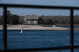 A small house with a rustic design sits on a narrow strip of land surrounded by water. The foreground features a steel railing, and the body of water is calm with a clear blue surface. Trees with lush foliage fill the background, while a buoy floats in the water and a small boat is docked near the shore.