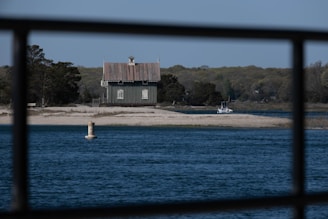 A small house with a rustic design sits on a narrow strip of land surrounded by water. The foreground features a steel railing, and the body of water is calm with a clear blue surface. Trees with lush foliage fill the background, while a buoy floats in the water and a small boat is docked near the shore.