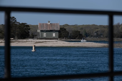 A small house with a rustic design sits on a narrow strip of land surrounded by water. The foreground features a steel railing, and the body of water is calm with a clear blue surface. Trees with lush foliage fill the background, while a buoy floats in the water and a small boat is docked near the shore.