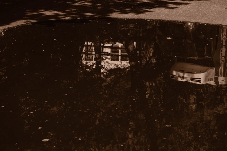 The image depicts a serene scene with a white car parked on a street, facing towards the right side of the frame and partially obscured by a tree reflection in the puddle below it. The background is a light brown color, providing a contrast to the white car and adding depth to the composition. The image has a vintage feel due to its black and white tones, giving it an artistic quality that draws the viewer's attention to the interplay of light and shadow.
