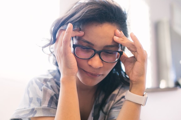 A person with long dark hair and glasses appears to be in a contemplative or stressed state, holding their temples with both hands. The background is softly blurred, giving a shallow depth of field effect, and the overall lighting is soft and natural.