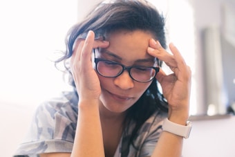 A person with long dark hair and glasses appears to be in a contemplative or stressed state, holding their temples with both hands. The background is softly blurred, giving a shallow depth of field effect, and the overall lighting is soft and natural.