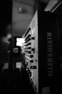 A close-up view of a sound mixing console with numerous dials and switches. The name 'ALLEN & HEATH' is visible on the side, indicating the brand of the equipment. The image is in black and white, adding a classic, dramatic feel to the scene.