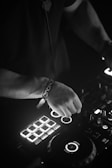 A person is operating a DJ controller with illuminated buttons and dials, indicating a dynamic and engaging music setup. The black and white color scheme adds a somber and focused atmosphere.