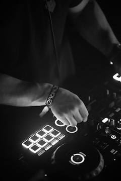 A person is operating a DJ controller with illuminated buttons and dials, indicating a dynamic and engaging music setup. The black and white color scheme adds a somber and focused atmosphere.