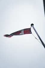 A Canadian flag is slightly tattered, waving from the top of a flagpole against an overcast sky. The flag's red and white colors are muted, and the maple leaf design is partially visible while the flag flutters.