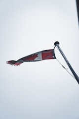 A Canadian flag is slightly tattered, waving from the top of a flagpole against an overcast sky. The flag's red and white colors are muted, and the maple leaf design is partially visible while the flag flutters.