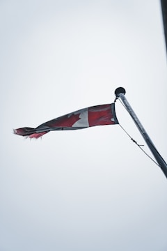 A Canadian flag is slightly tattered, waving from the top of a flagpole against an overcast sky. The flag's red and white colors are muted, and the maple leaf design is partially visible while the flag flutters.