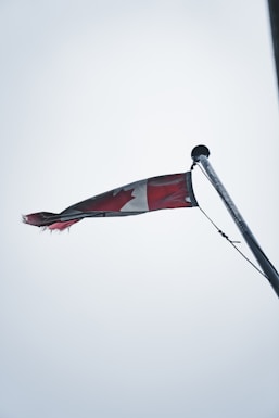 A Canadian flag is slightly tattered, waving from the top of a flagpole against an overcast sky. The flag's red and white colors are muted, and the maple leaf design is partially visible while the flag flutters.