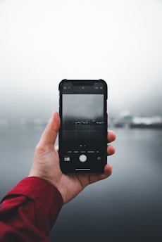 A hand wearing a red sleeve holds a smartphone in an outdoor setting. The phone screen displays a gloomy, foggy landscape with water and indistinct distant objects.