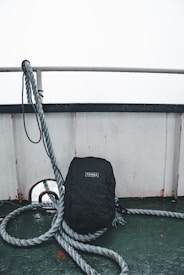 A black backpack with a 'Tenba' logo is leaning against a ship's railing. Thick ropes are coiled around the backpack, and the deck surface is a worn, greenish color. The top part of the image is dominated by an overcast sky and the stark, white railing.