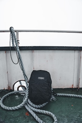 A black backpack with a 'Tenba' logo is leaning against a ship's railing. Thick ropes are coiled around the backpack, and the deck surface is a worn, greenish color. The top part of the image is dominated by an overcast sky and the stark, white railing.