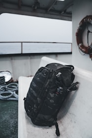 A black camouflage backpack is resting on a white bench aboard a ship. The background shows a lifebuoy and coiled ropes, with the sea visible through the windows.
