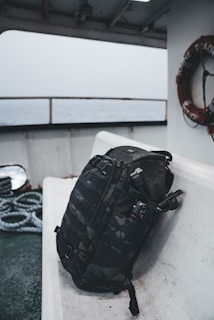 A black camouflage backpack is resting on a white bench aboard a ship. The background shows a lifebuoy and coiled ropes, with the sea visible through the windows.