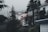 A foggy residential area with multiple houses surrounded by tall evergreen trees. The houses are mostly wooden with some white siding, and one has a small elevated deck. The fog adds a misty atmosphere to the scene.