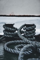 Thick ropes are tightly wound around two metal posts on a ship deck, with a background of the sea visible. The scene has a rugged, maritime feel.