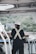 A person wearing a work suit and a white helmet stands on a boat, looking out over the water. The individual is positioned near ropes and metal railings, with a natural landscape in the background featuring greenery and hazy outlines of distant structures.