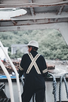 A person wearing a work suit and a white helmet stands on a boat, looking out over the water. The individual is positioned near ropes and metal railings, with a natural landscape in the background featuring greenery and hazy outlines of distant structures.