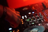 A close-up of a DJ's hand operating a mixing console. The mixer is surrounded by various knobs and buttons, with colored lights illuminating the surface. The scene is bathed in warm red lighting, creating a vibrant and dynamic atmosphere.