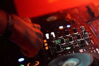 A close-up of a DJ's hand operating a mixing console. The mixer is surrounded by various knobs and buttons, with colored lights illuminating the surface. The scene is bathed in warm red lighting, creating a vibrant and dynamic atmosphere.