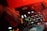 A close-up of a DJ's hand operating a mixing console. The mixer is surrounded by various knobs and buttons, with colored lights illuminating the surface. The scene is bathed in warm red lighting, creating a vibrant and dynamic atmosphere.