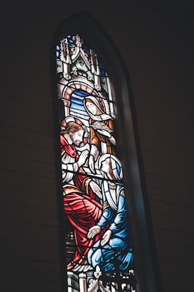 A stained glass window featuring a biblical scene with three figures. The central figure is depicted with a halo and red robes, while another figure kneels, dressed in blue with a white head covering. The third figure, standing, wears orange-brown garments and holds a bowl. The background shows architectural details and vibrant colors.
