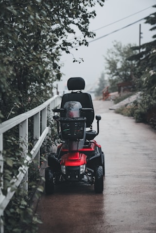 A red mobility scooter is parked on a wet, paved pathway, surrounded by greenery. The scene appears to be outdoors, with a tall wooden fence lining the path and trees overhead.