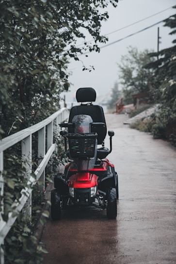 A red mobility scooter is parked on a wet, paved pathway, surrounded by greenery. The scene appears to be outdoors, with a tall wooden fence lining the path and trees overhead.