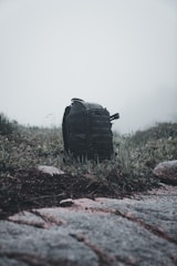 A black backpack sits on a patch of grass surrounded by rocks and weeds under a foggy, overcast sky. The scene appears to be in a natural, rugged outdoor setting, possibly suggesting a hiking trip or a camping adventure.