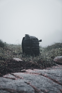 A black backpack sits on a patch of grass surrounded by rocks and weeds under a foggy, overcast sky. The scene appears to be in a natural, rugged outdoor setting, possibly suggesting a hiking trip or a camping adventure.