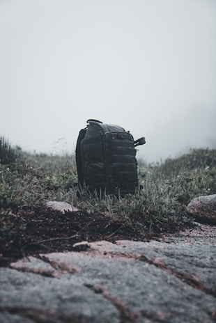 A black backpack sits on a patch of grass surrounded by rocks and weeds under a foggy, overcast sky. The scene appears to be in a natural, rugged outdoor setting, possibly suggesting a hiking trip or a camping adventure.