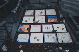 A collection of decorative tiles laid out in a grid, each featuring unique designs such as a guitar, a unicorn, abstract patterns, leaves, and inscribed messages. They are surrounded by small decorative objects and figurines, including a black swan sculpture. The setting appears outdoors with a wooden deck area.