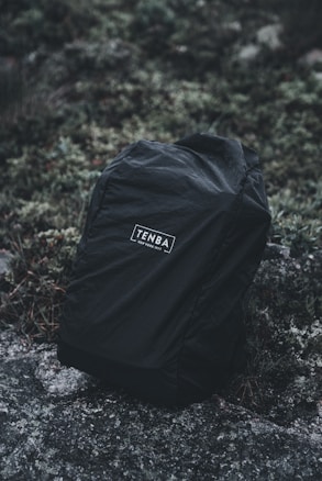 A black backpack with the brand name 'TENBA' printed on the front, placed on a natural, rocky surface surrounded by moss and greenery.