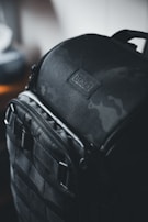 A rugged black backpack with a sleek design, featuring a slightly reflective surface and sturdy straps. The bag has a label that reads 'Tenba' with 'New York' inscribed below it. The lighting is dim, creating a moody and sophisticated atmosphere.