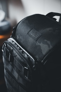 A rugged black backpack with a sleek design, featuring a slightly reflective surface and sturdy straps. The bag has a label that reads 'Tenba' with 'New York' inscribed below it. The lighting is dim, creating a moody and sophisticated atmosphere.